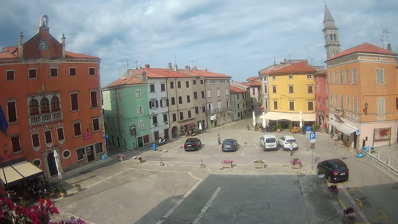 A town square with several colorful buildings, some cars parked, and a few people walking around under partly cloudy skies.