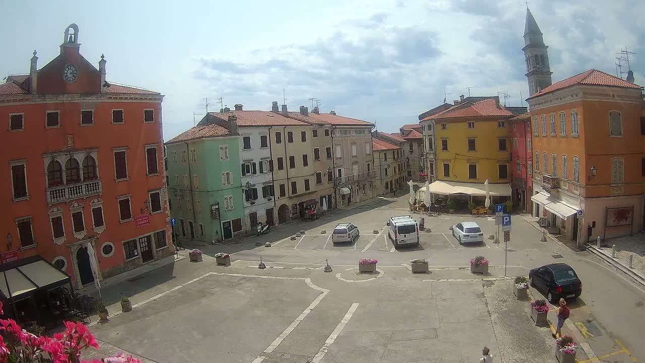A mostly sunny town square with several parked cars, colorful buildings, and a few pedestrians.