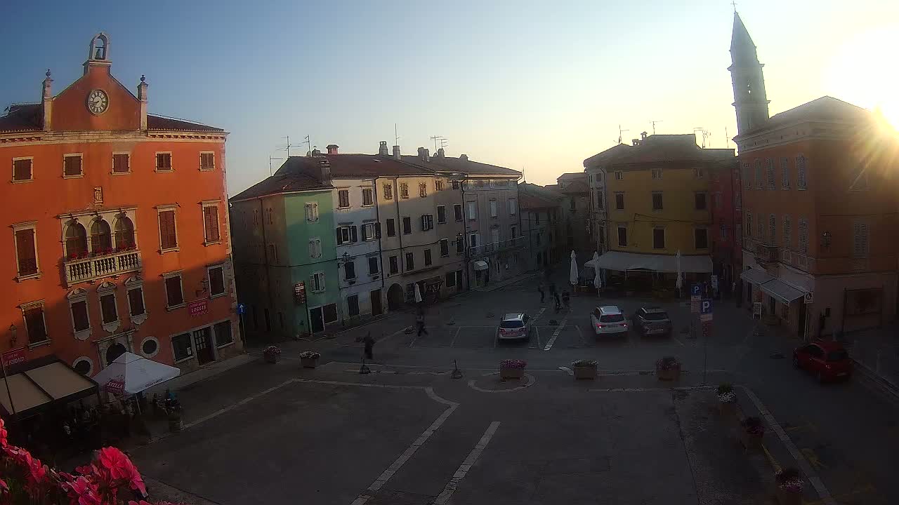 A town square with several multi-story buildings, some cars parked, and a few people walking around, all under a clear, sunny sky.