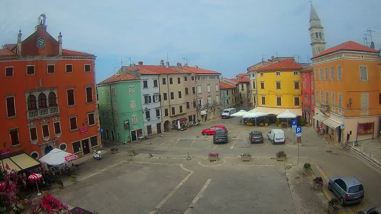 A town square with colorful buildings, some cars parked, and outdoor seating under umbrellas, on a cloudy day.