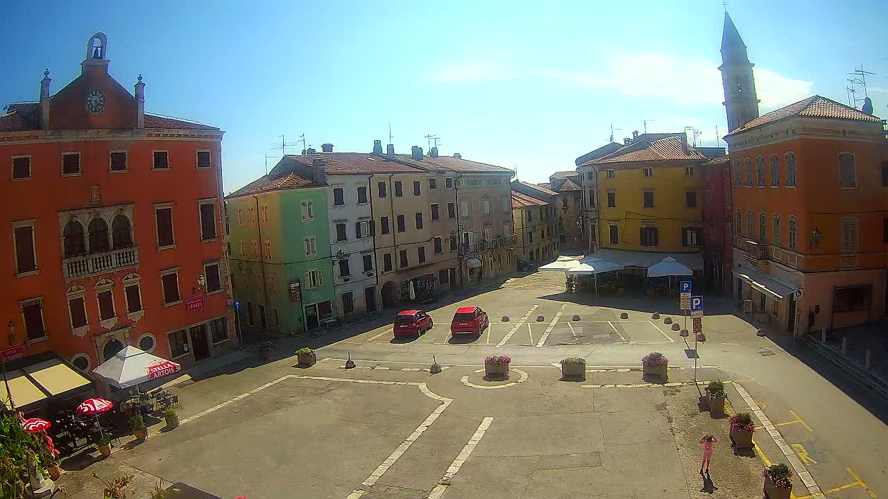 A sunny town square with colorful buildings, a few parked cars, outdoor seating areas, and a person in red walking across the pavement.