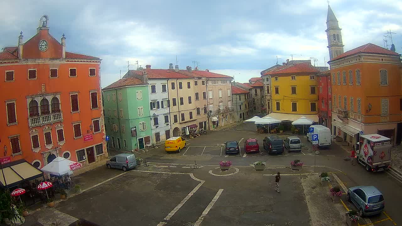 A town square with colorful buildings under a cloudy sky, containing parked cars and a few people.