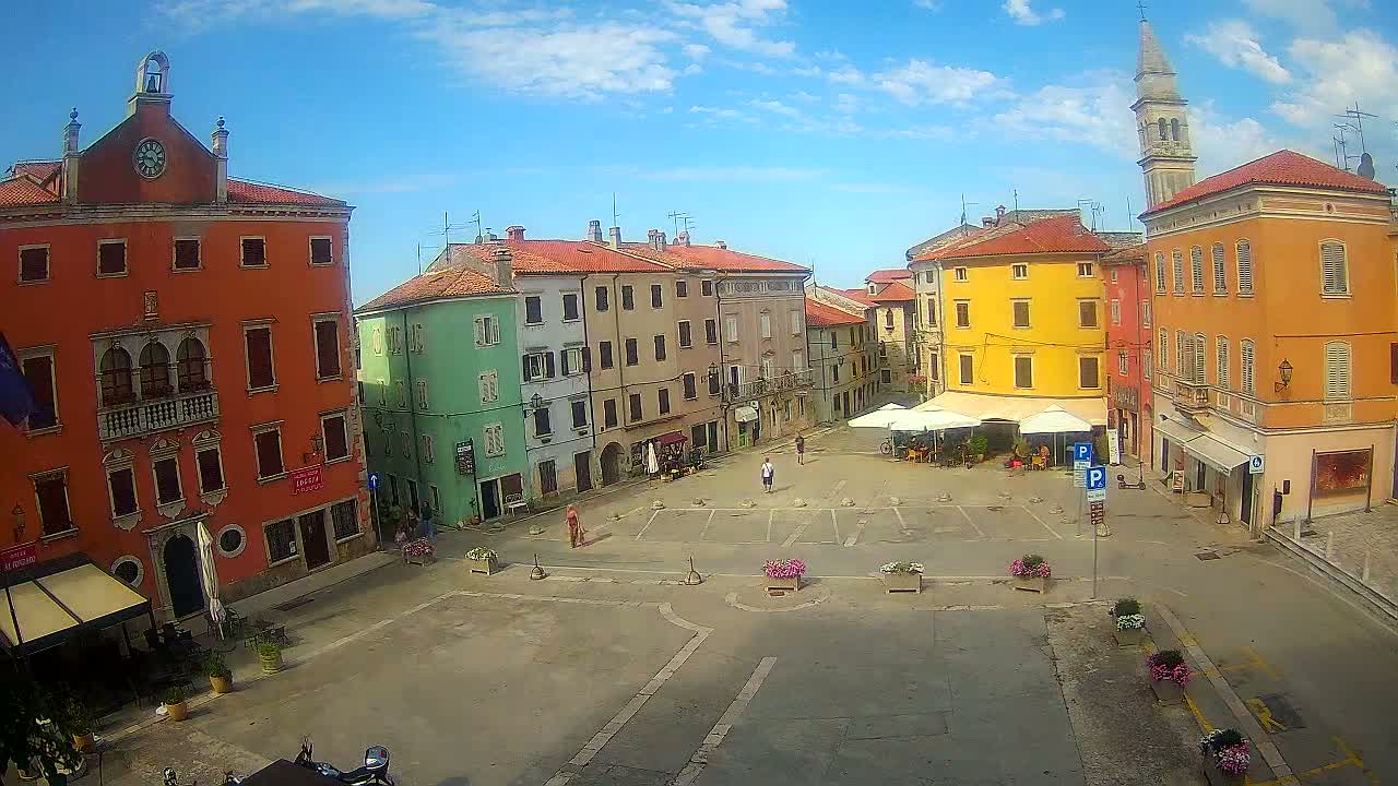 A sunny day reveals a town square surrounded by colorful buildings, some with awnings, and a few people are scattered about.