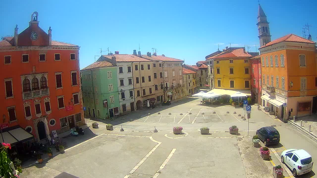 A sunny day reveals a town square surrounded by colorful buildings, some with awnings, and a few people are scattered about.