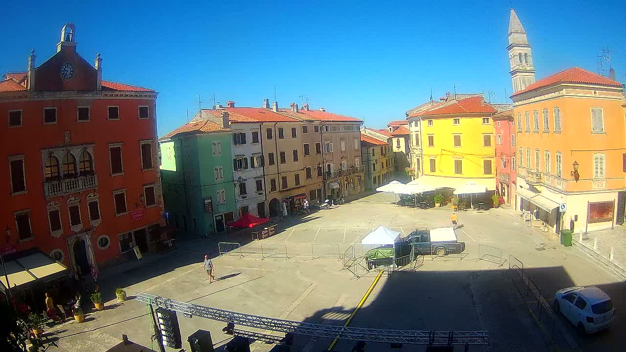 A town square with colorful buildings under a clear blue sky is partially occupied by temporary fencing, canopies, and a few vehicles.