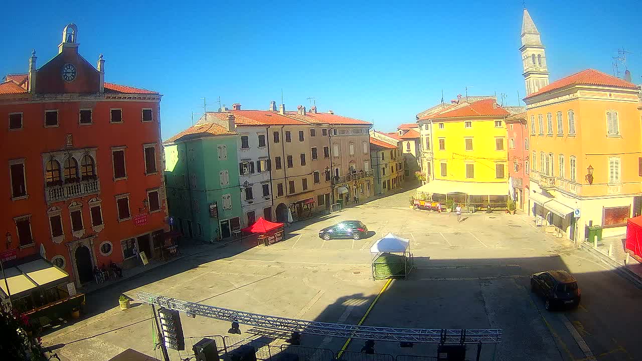 A sunny day reveals a town square surrounded by colorful, multi-story buildings, with a few cars and some outdoor seating visible.