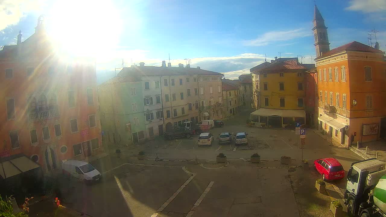 A sunny day reveals a town square surrounded by colorful, multi-story buildings, with a few cars and some outdoor seating visible.