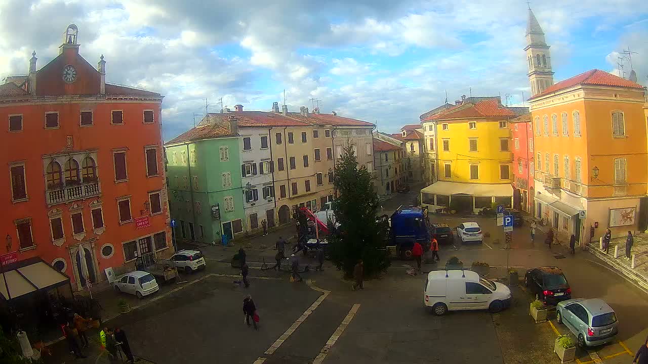 A vibrant town square is seen on a partly cloudy day, featuring historic buildings in various colors, pedestrians, parked cars, a large evergreen tree, and a truck with a crane, all overlooked by a distant church spire.