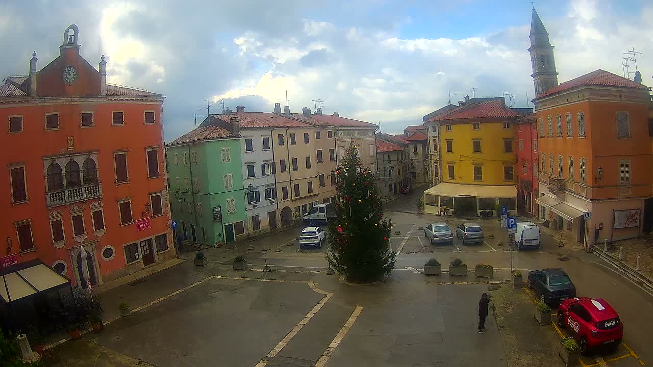 A vibrant town square features a decorated Christmas tree, a mix of colorful historic buildings including an orange one with a clock and a distant church bell tower, with several parked cars and a few pedestrians, under an overcast sky with patches of blue.