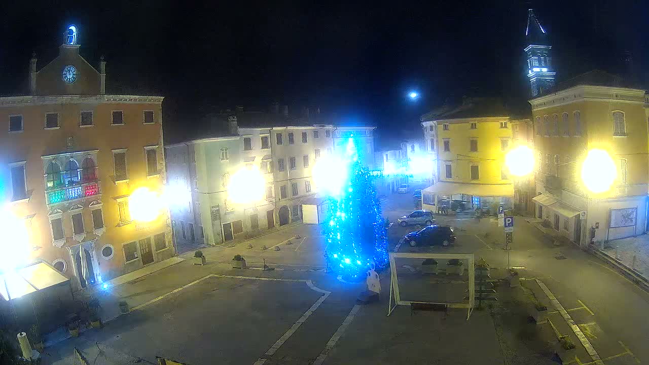 A clear night reveals a lively European-style town square with a brightly lit blue Christmas tree at its center, surrounded by historic buildings glowing with warm lights, parked cars, and a distant church tower under a visible moon.
