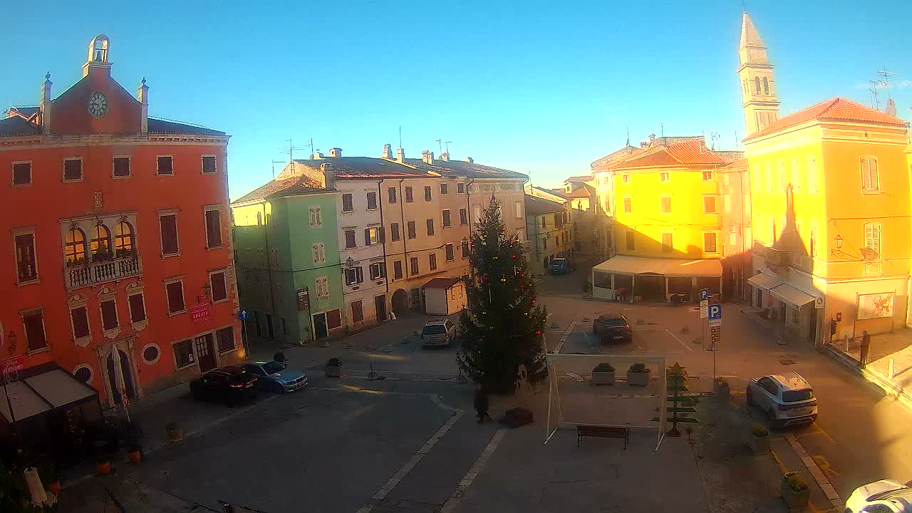 A clear blue sky and bright sun illuminate a picturesque European town square with colorful historic buildings, a tall church steeple, a decorated Christmas tree, and scattered parked cars and pedestrians.