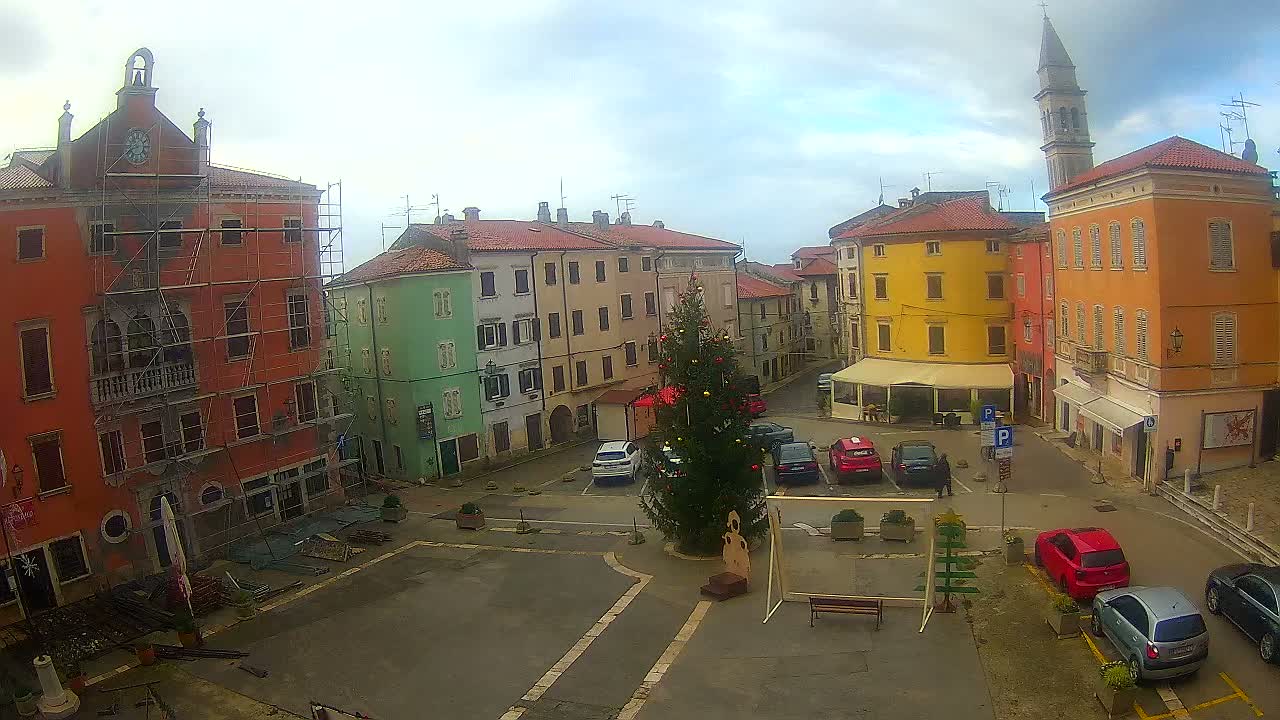 A vibrant town square features a decorated Christmas tree and parked cars, surrounded by colorful historic buildings—one with scaffolding—all under an overcast sky with a bell tower visible in the distance.
