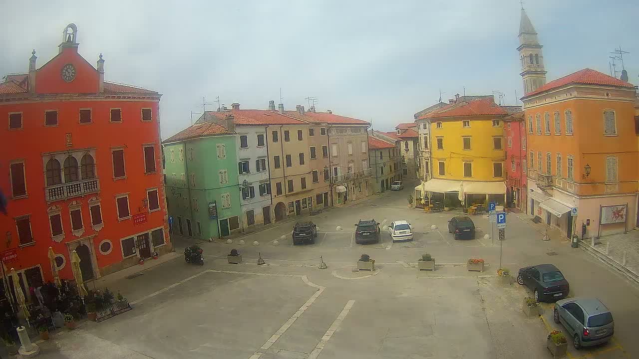 A vibrant town square features a decorated Christmas tree and parked cars, surrounded by colorful historic buildings—one with scaffolding—all under an overcast sky with a bell tower visible in the distance.