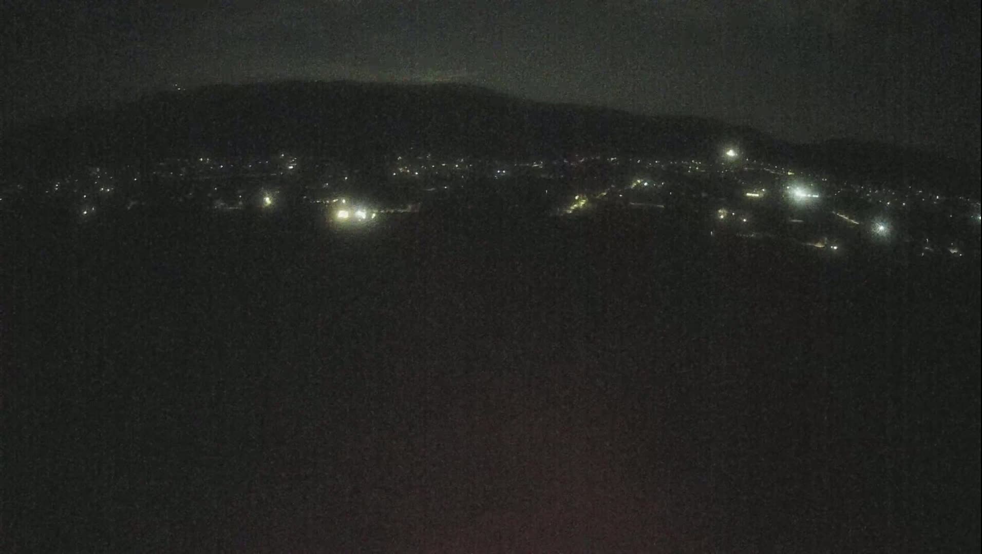 A nighttime long shot shows a dark landscape with a few scattered lights of a town below a dark mountain range under a clear, dark sky.