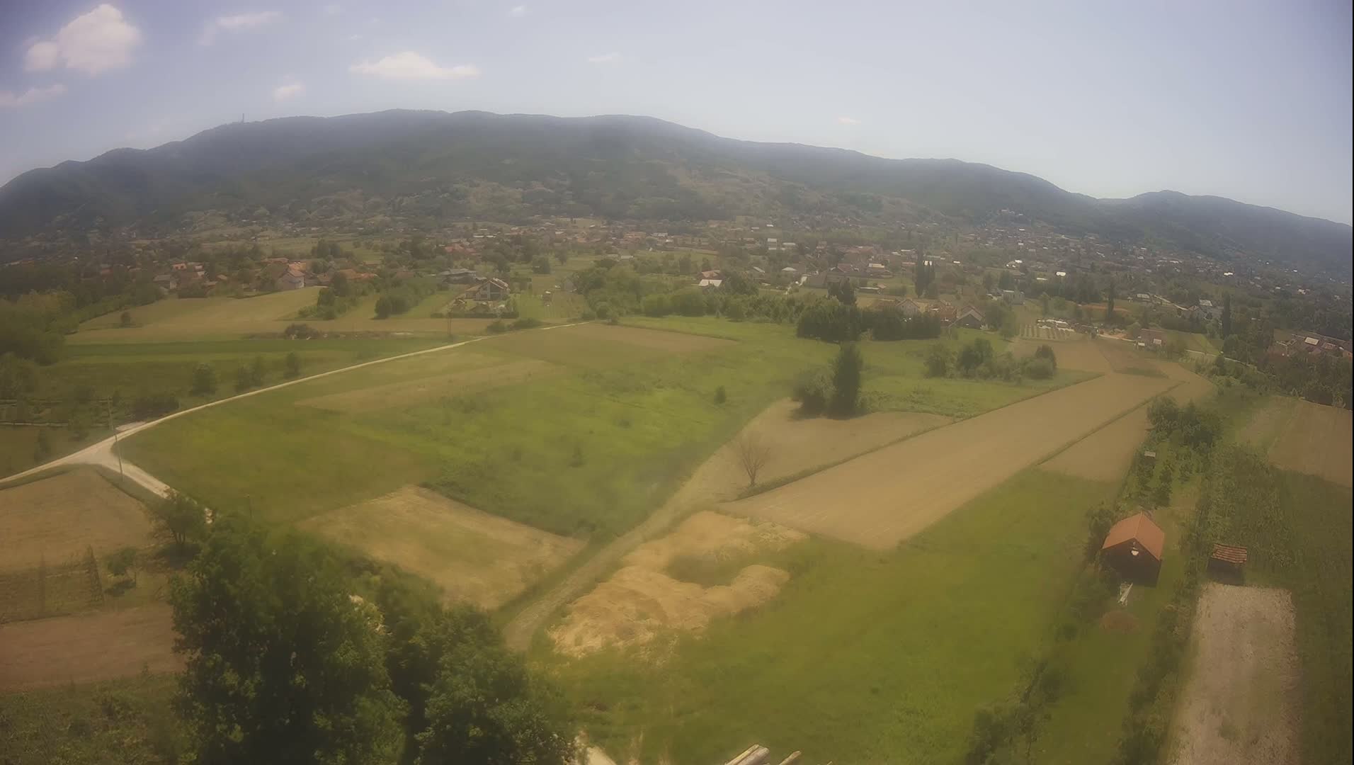 An aerial view shows a rural landscape under a partly sunny sky, featuring farmland divided into sections, a small village nestled in a valley, and distant mountains.