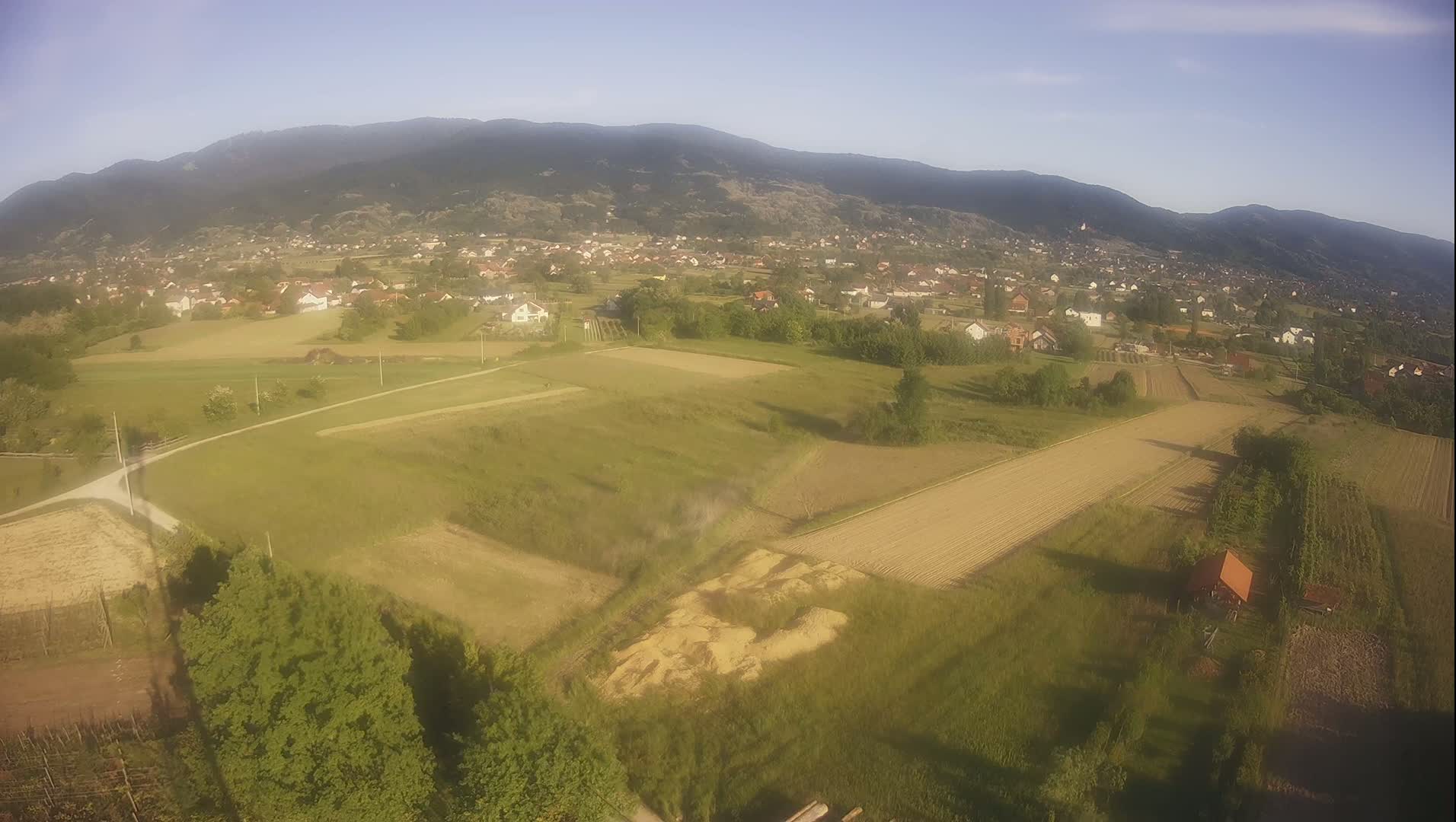 An aerial view shows a rural landscape with cultivated fields, scattered houses, and a mountain range in the background under a clear, bright sky.
