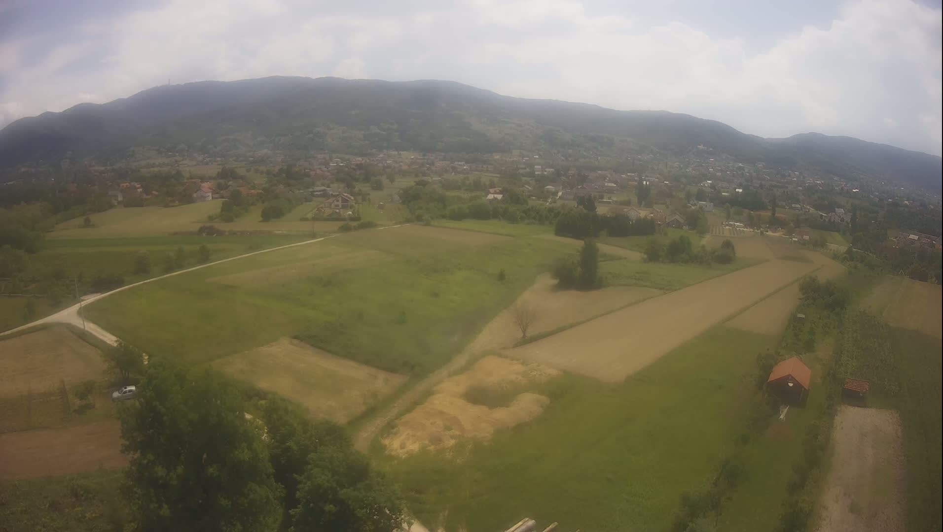 An aerial view shows a rural landscape under a partly cloudy sky, featuring cultivated fields, a small village nestled in a valley, and a distant mountain range.