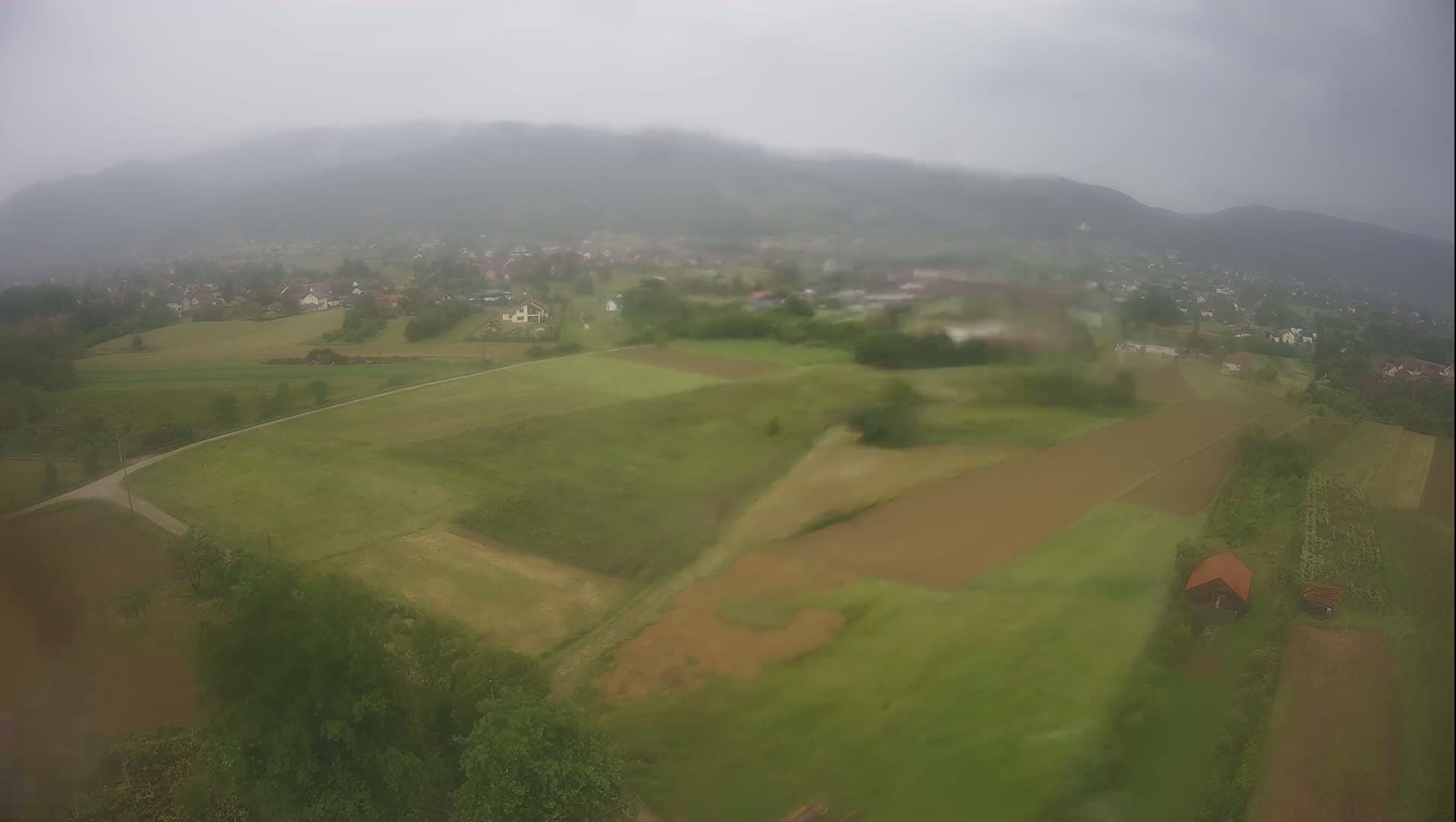 An aerial view shows a rain-soaked rural landscape with farmlands and a small village nestled among rolling hills under a cloudy sky.