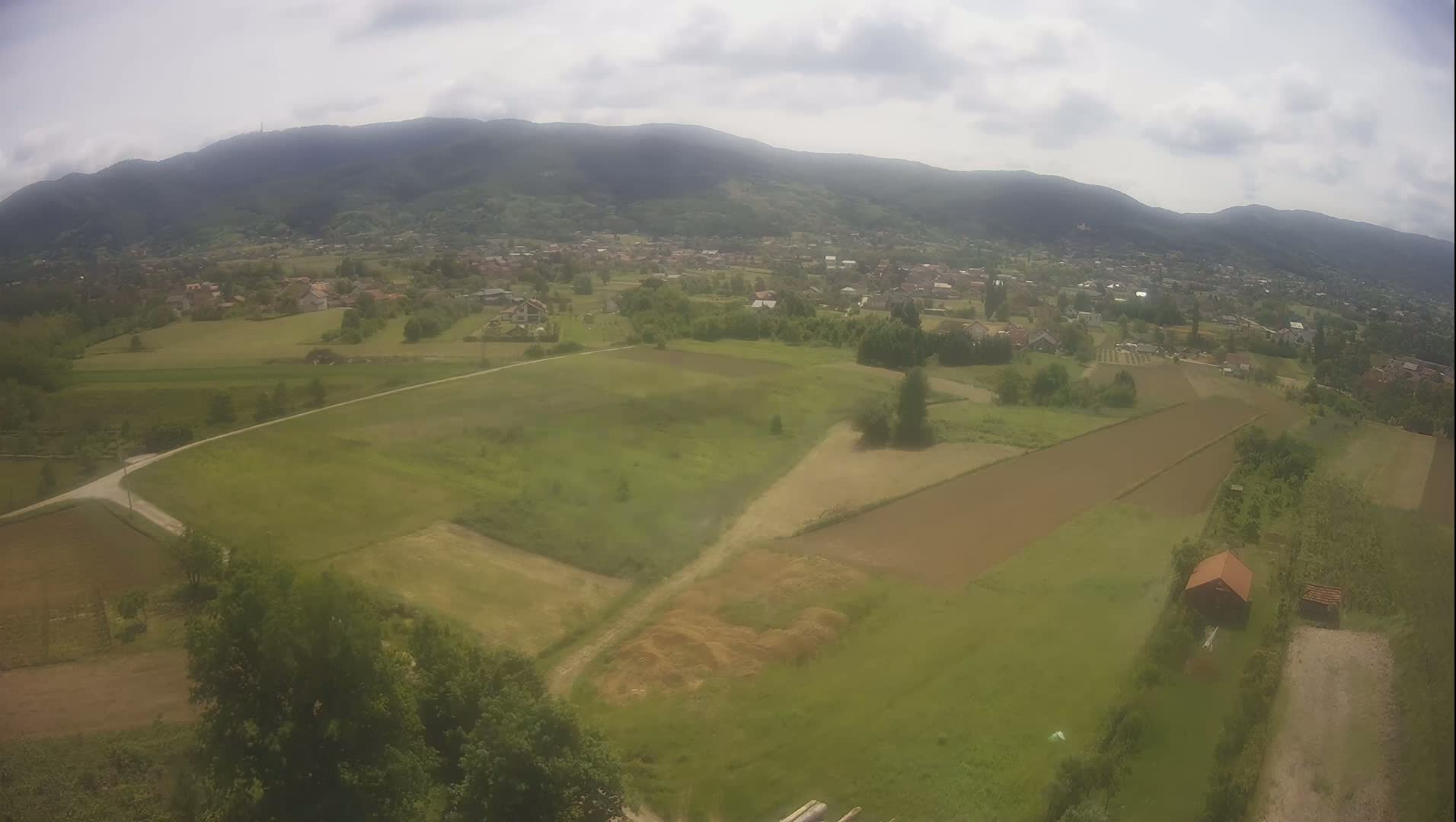 An aerial view shows a rural landscape with green fields, a small village nestled in a valley, and mountains in the background under a partly cloudy sky.