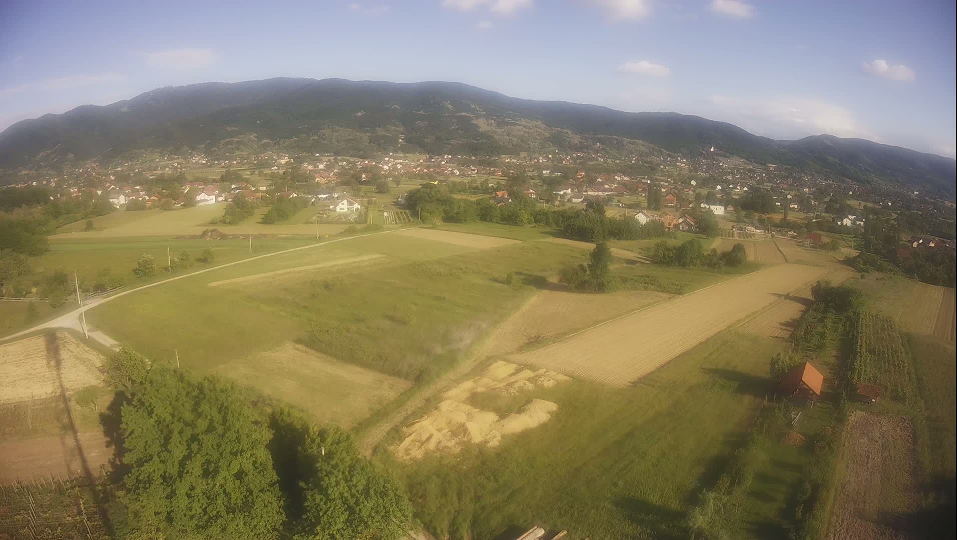 An aerial view shows a rural landscape of fields, a small village, and mountains under a mostly clear, sunny sky.