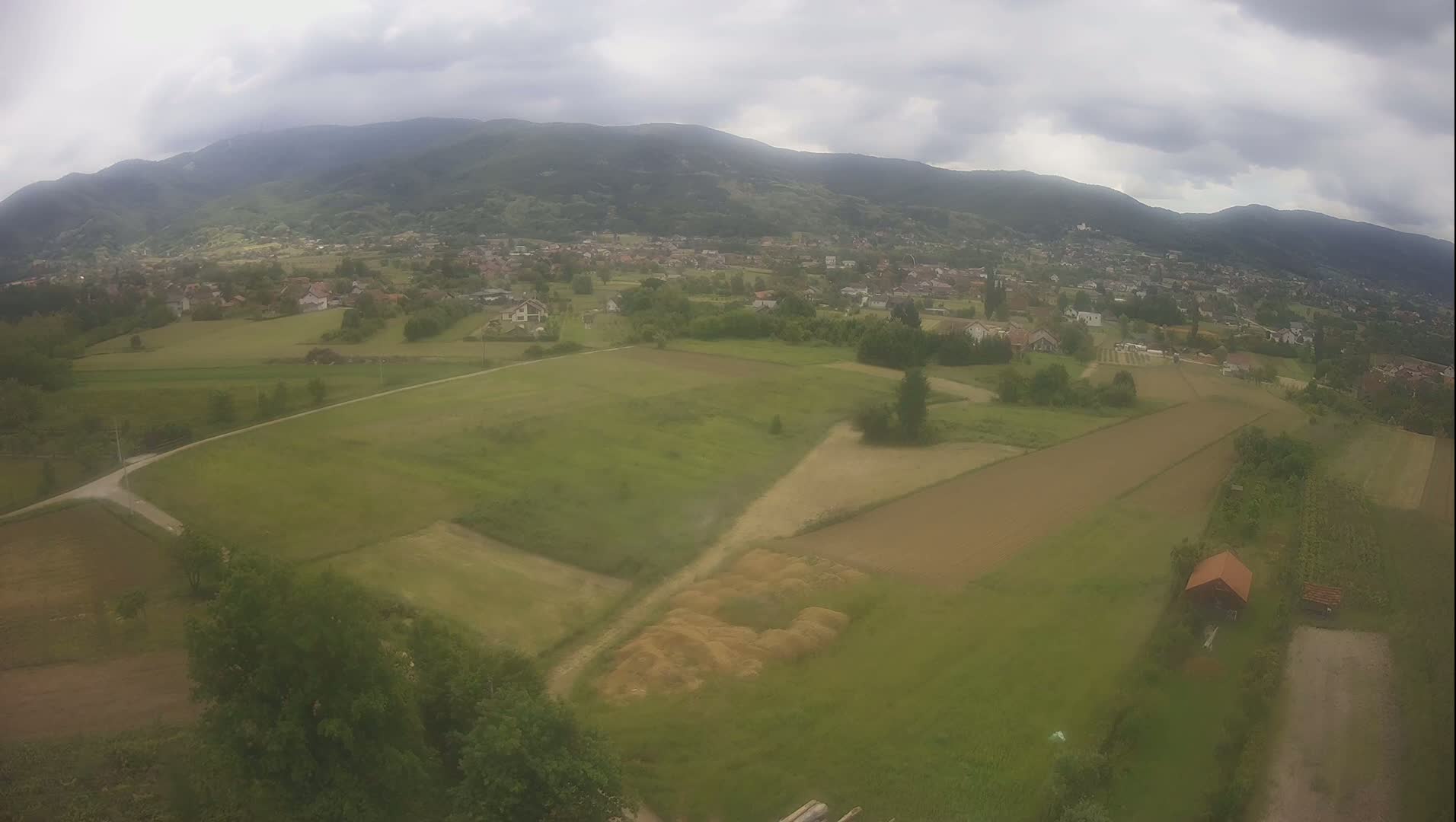 An aerial view shows a rural landscape with farmlands, a small village nestled in a valley, and mountains in the background under an overcast sky.