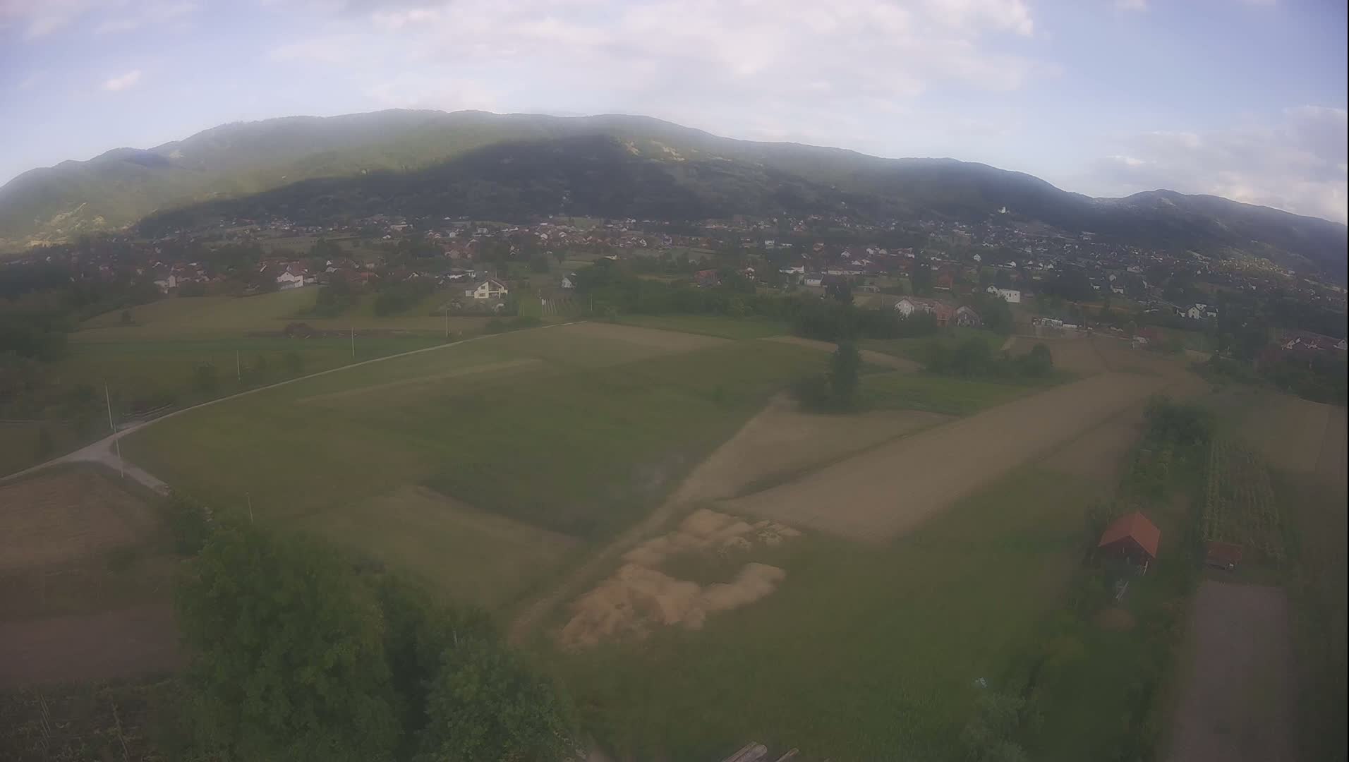 An aerial view shows a small village nestled in a valley, surrounded by farmland and a hazy mountain range under a partly cloudy sky.