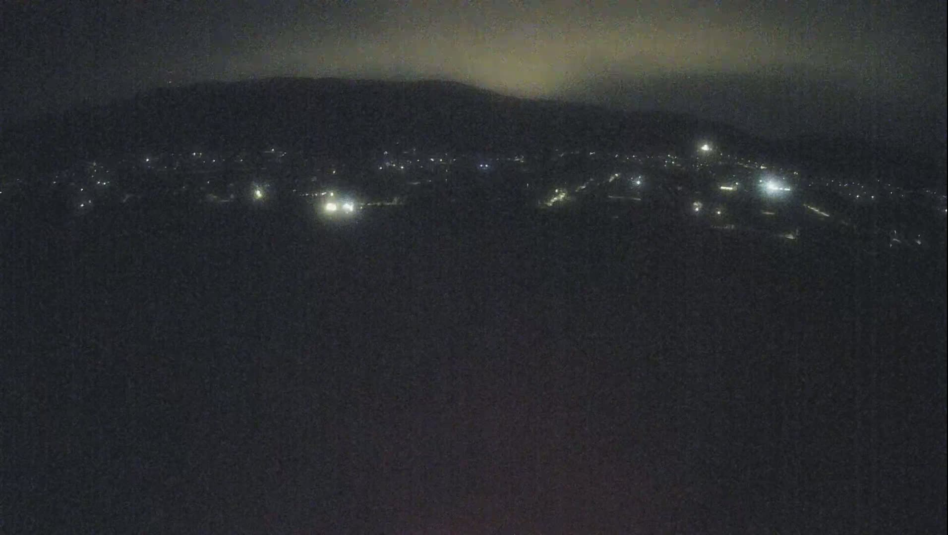 A nighttime, long shot shows a dimly lit town nestled below a dark mountain range under a cloudy sky.