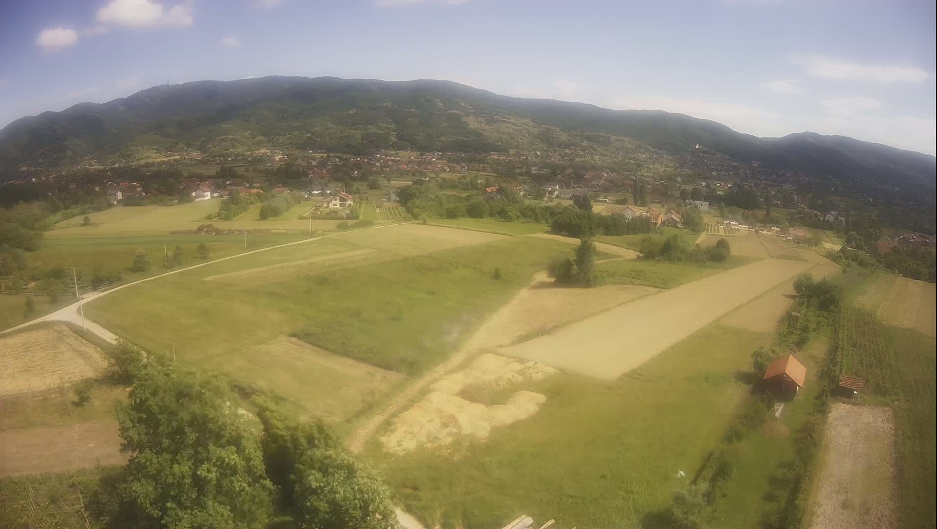 An aerial view shows a rural landscape under a partly sunny sky, with green fields, a small village nestled in a valley, and mountains in the background.