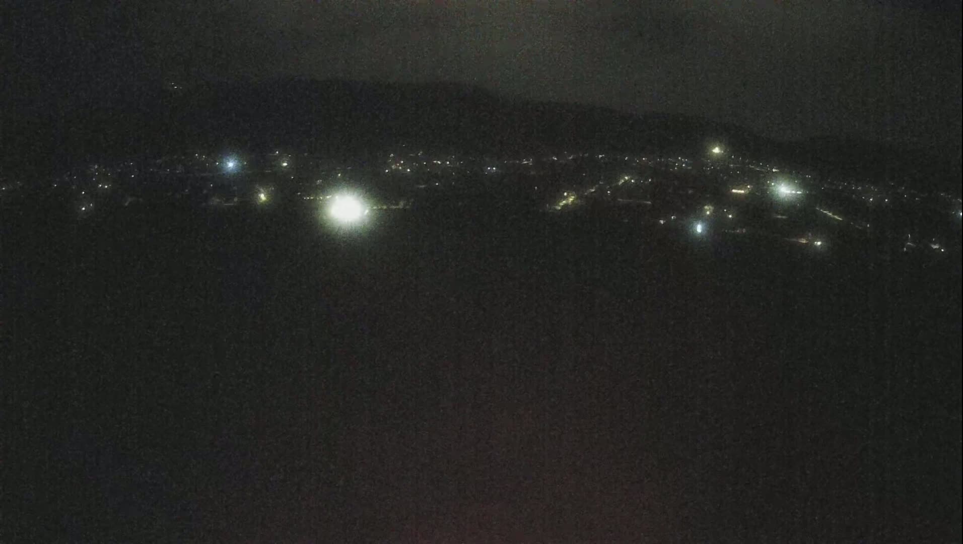 A nighttime, long-range view shows a dark landscape dotted with sparsely scattered lights from a town or village, partially obscured by a dark sky.