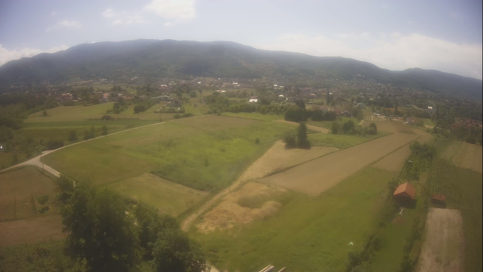 An aerial view shows a rural landscape under a partly sunny sky, with green fields, a small village nestled among the fields, and a range of mountains in the background.