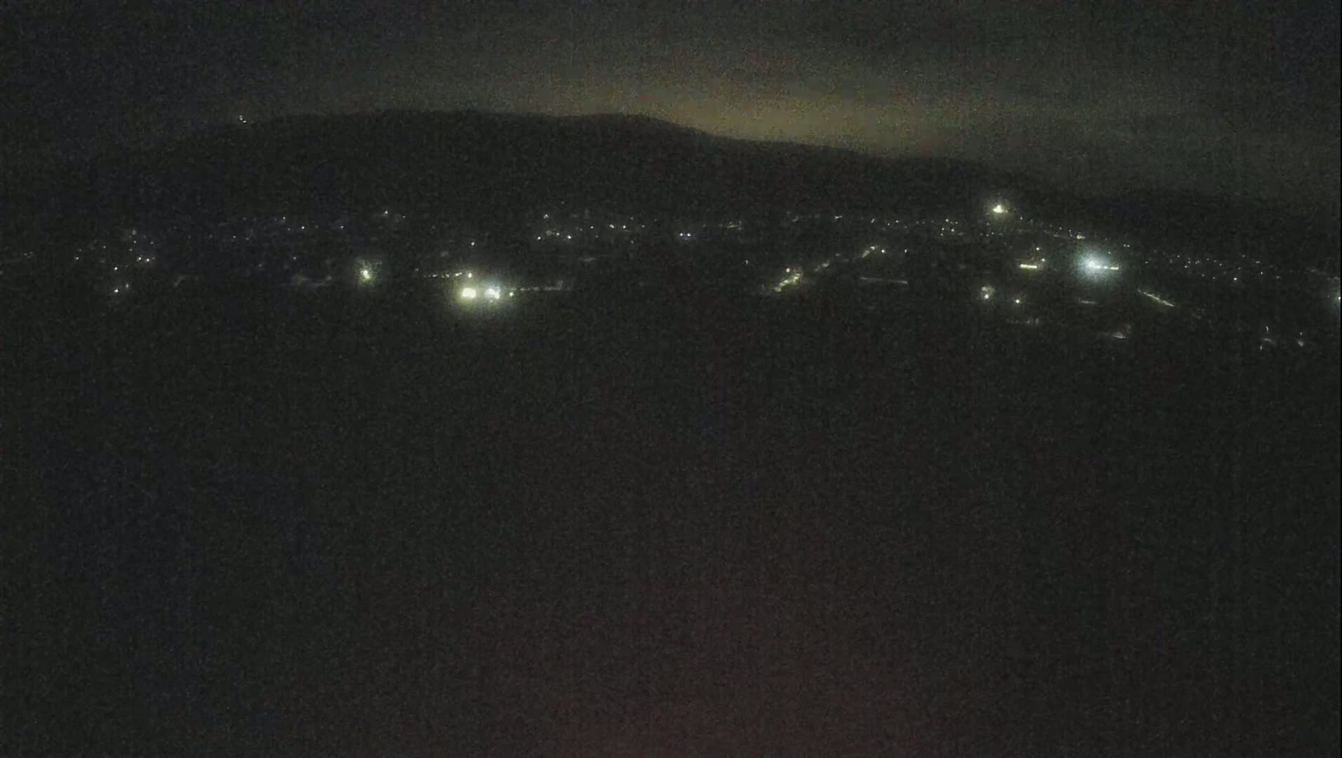 A nighttime, long shot of a town nestled below a dark mountain range under a cloudy sky.