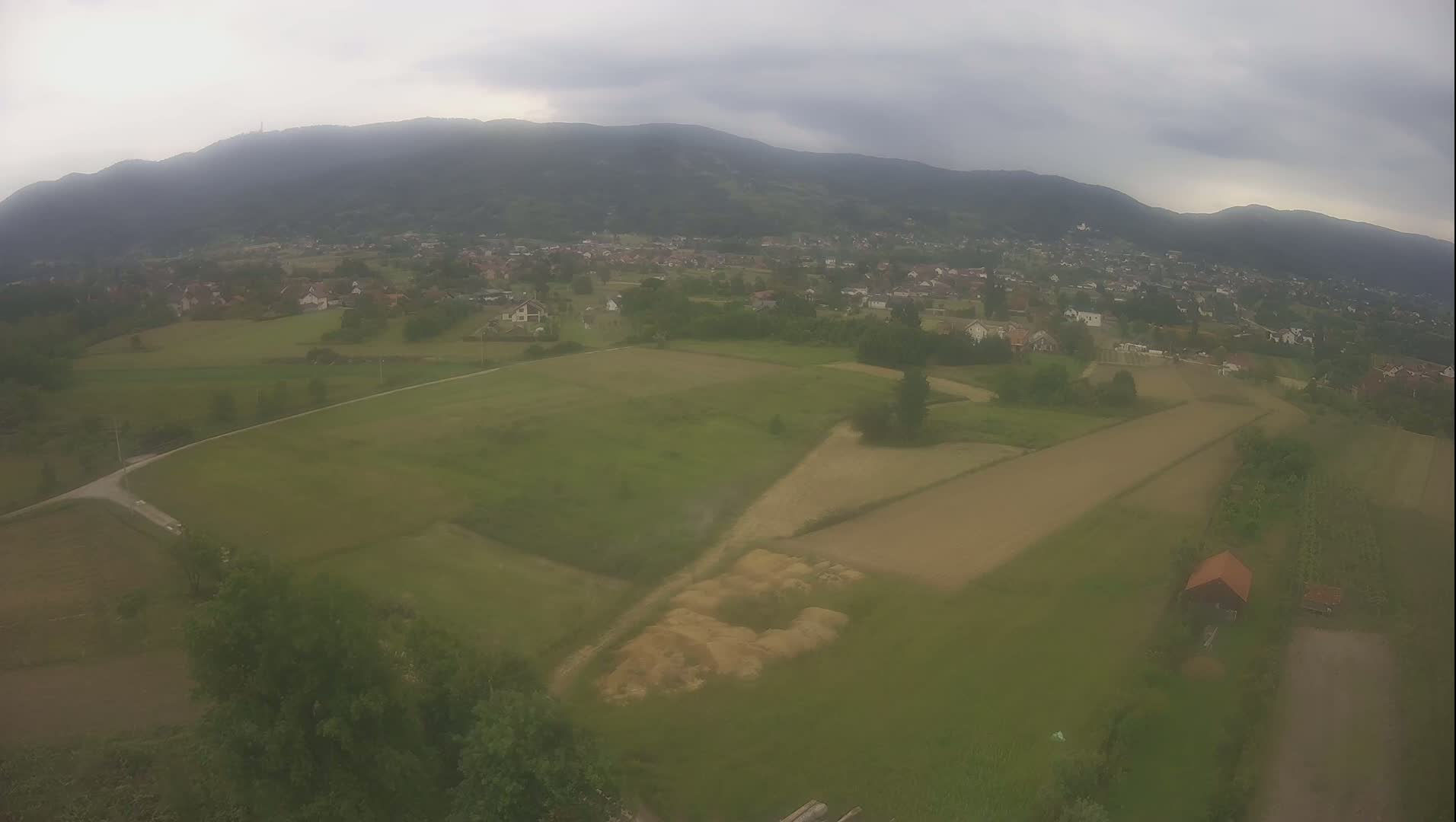 An aerial view shows a hazy, overcast day over a rural landscape of green fields, a small village, and distant mountains.