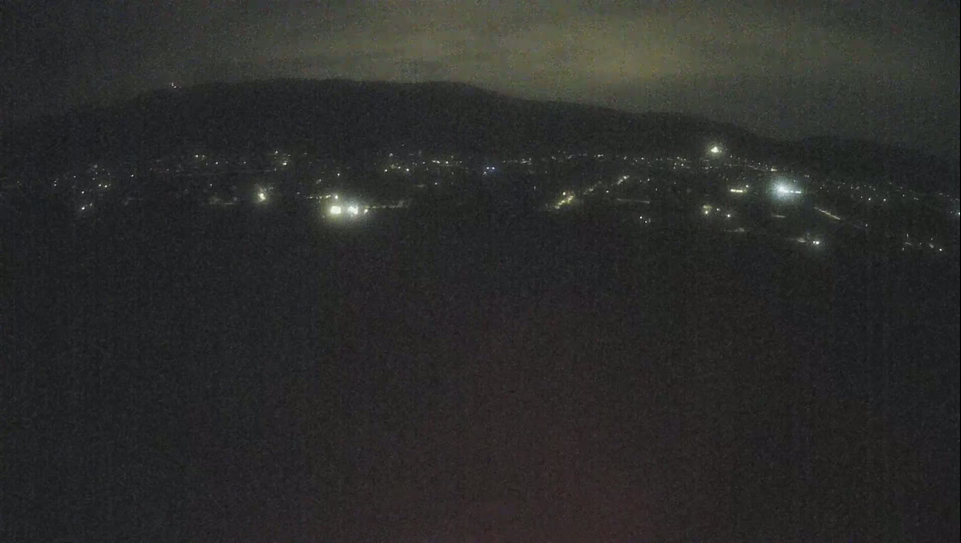 A nighttime, long shot shows a dark landscape with a faintly lit town nestled below a dark mountain range under a cloudy sky.