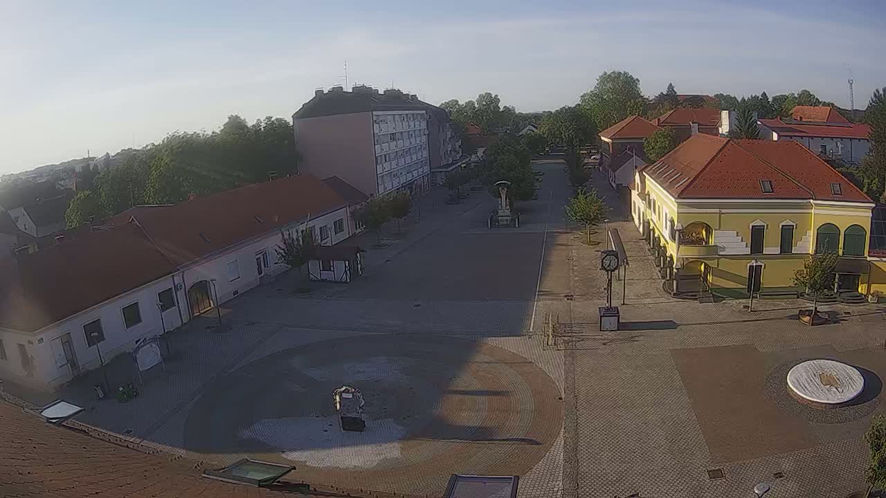 A town square with several buildings surrounding a paved central area, under a clear, sunny sky.