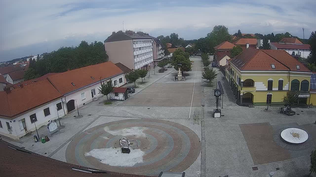 A town square with several buildings surrounding a paved area featuring a circular mosaic design, under partly cloudy skies.