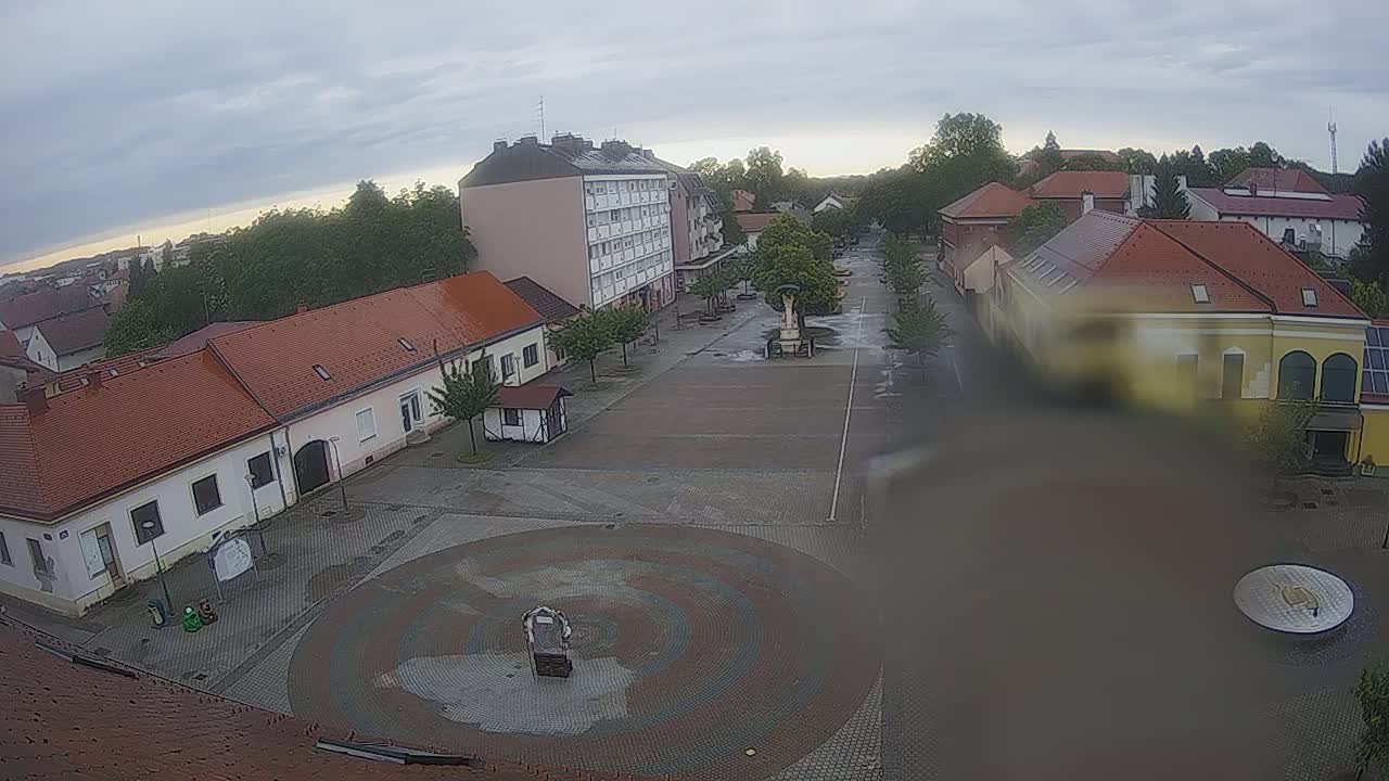 A town square with several low-rise buildings and a circular paved area in the center, under an overcast sky.