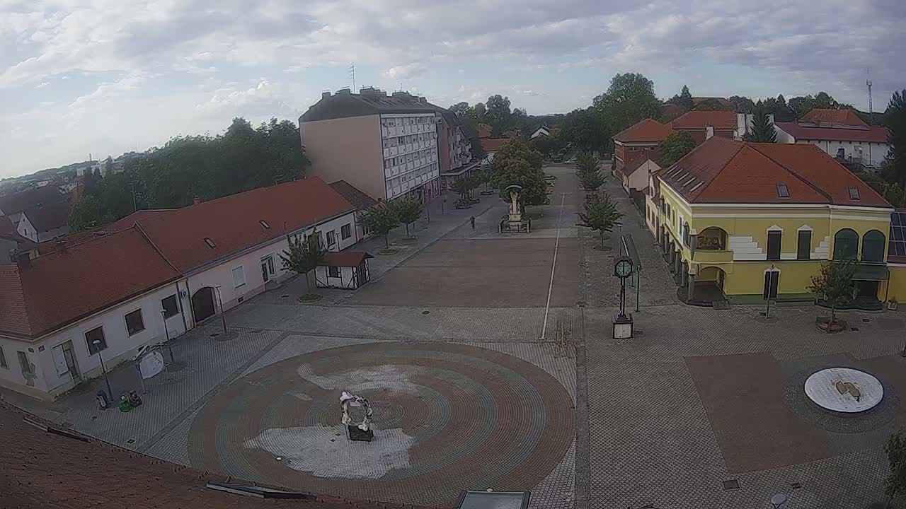 A mostly cloudy day reveals a town square with low buildings and a central circular paved area, surrounded by trees.