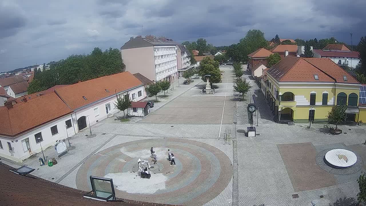 A town square with several buildings surrounding a paved area containing a circular fountain and a few people, under a partly cloudy sky.