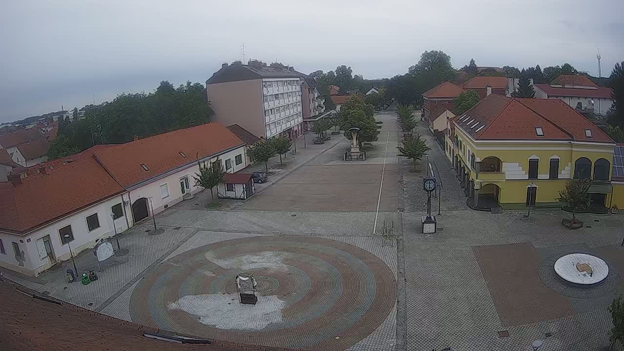 A town square with low buildings and a large circular paved area is seen under an overcast sky.