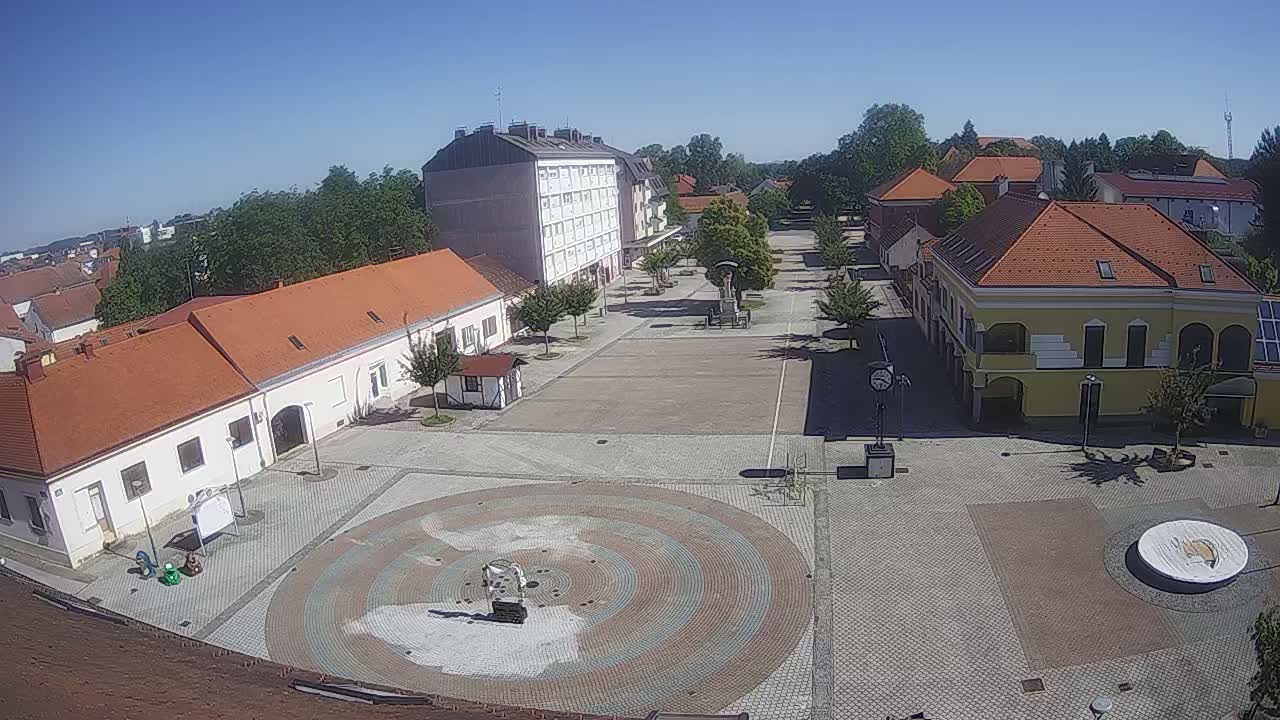A town square with several buildings surrounding a paved central area featuring a circular, multi-colored design, under a clear, sunny sky.