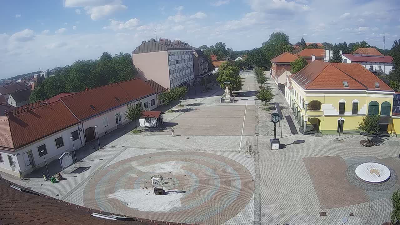 A mostly empty town square with low buildings and red tile roofs, under a partly cloudy sky.