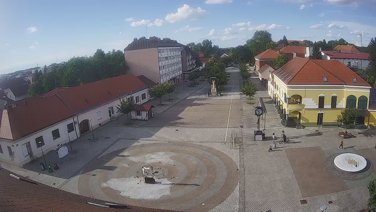 A town square with several low buildings under a mostly sunny sky, featuring a circular paved area in the center.