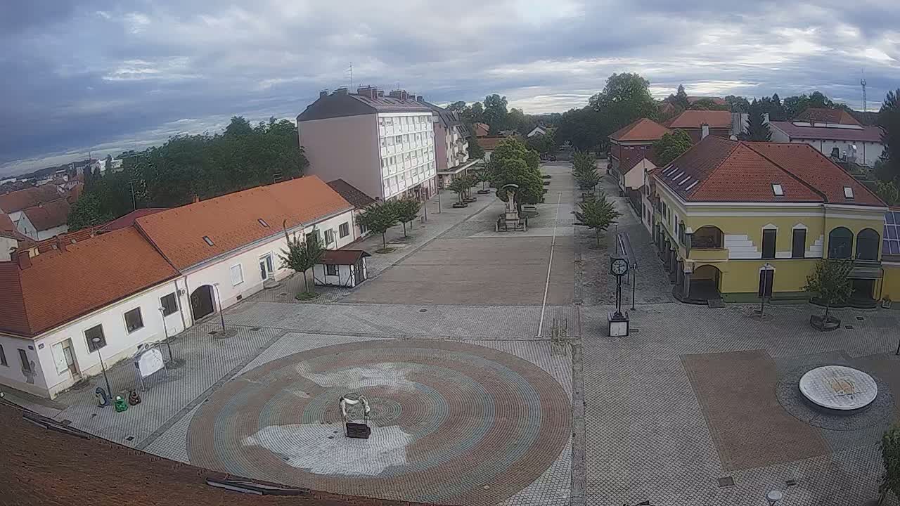 A mostly empty town square with low buildings and trees under a cloudy sky.