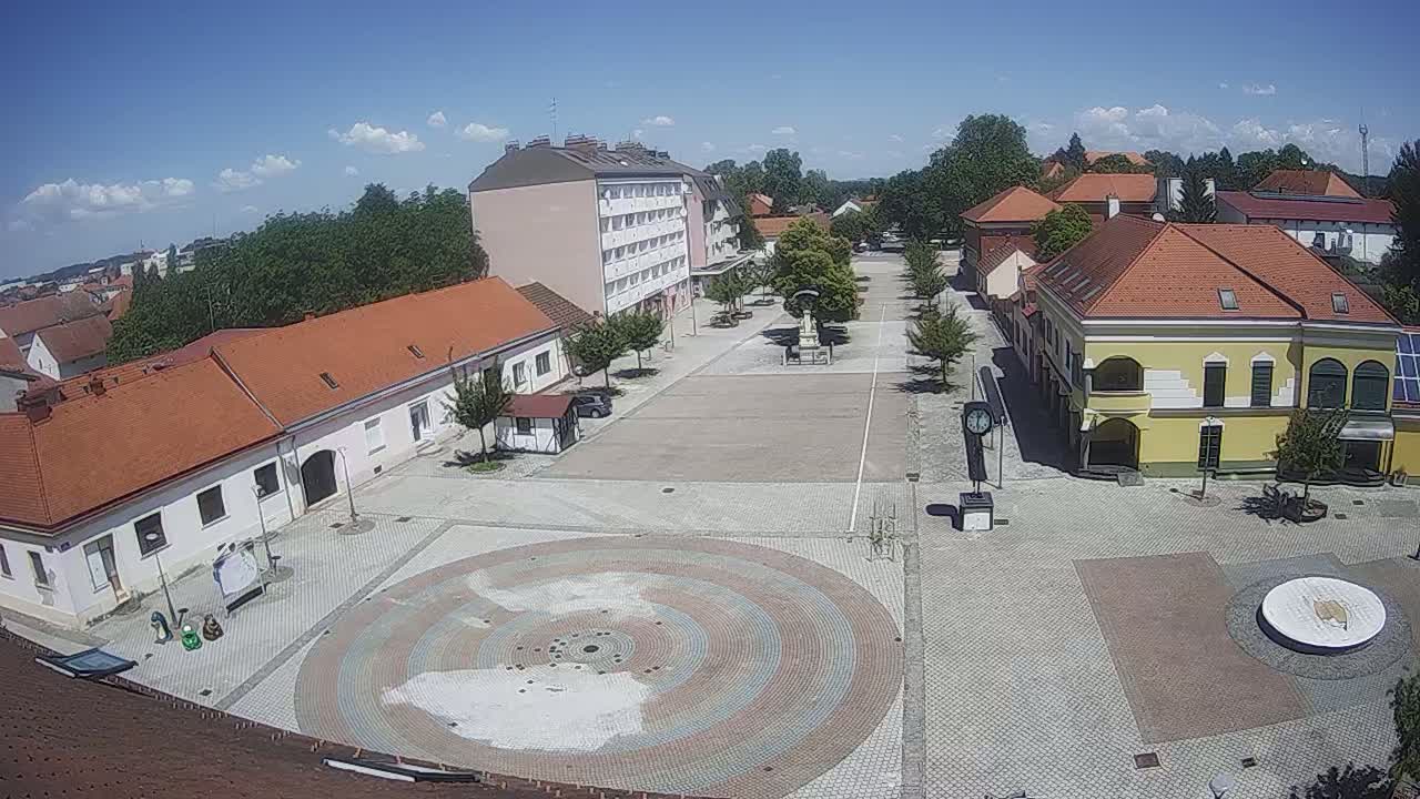 A town square with low buildings and a circular paved area in the center, under sunny skies with a few clouds.