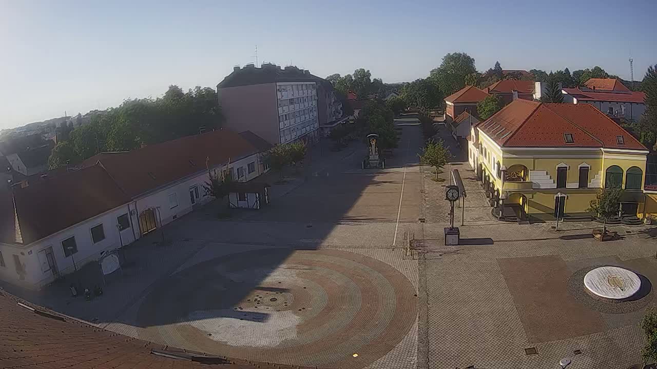 A sunny day reveals a town square with several buildings surrounding a circular paved area, featuring a tall building and a yellow building with arched entryways.