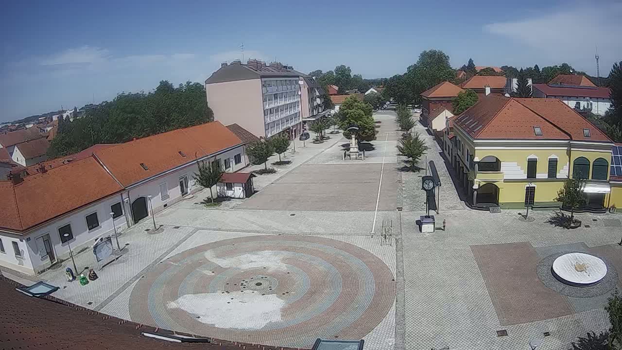 A town square with several low buildings surrounding a paved central area featuring a circular mosaic design, all under a clear sunny sky.