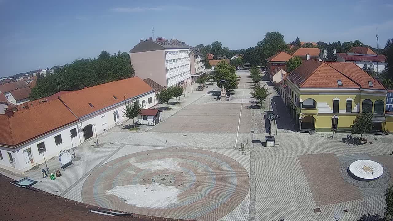 A large town square with buildings surrounding it, under sunny skies, features a circular paved area in the center.