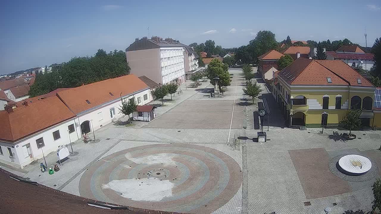 A town square with several buildings surrounding a paved area featuring a circular, multi-colored design in the center, all under a clear, sunny sky.
