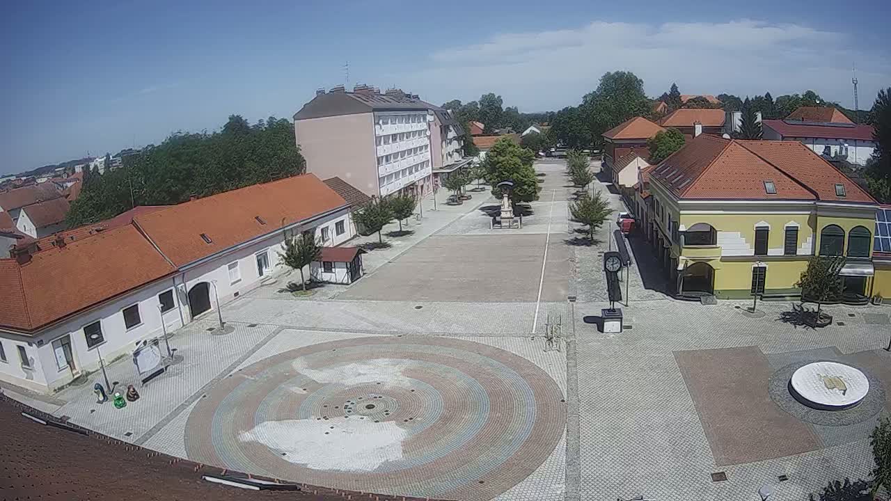 A town square with several buildings, trees, and a circular paved design in the center on a sunny day.