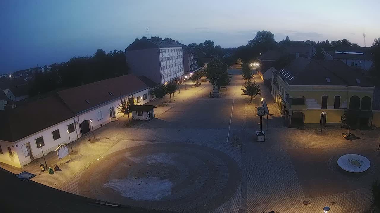 An illuminated town square at dusk, with low-rise buildings surrounding a central paved area containing a circular feature.